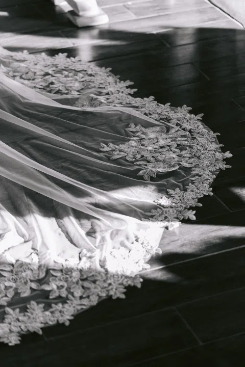 Detalle en blanco y negro del bordado y la cola de un vestido blanco extendido sobre el suelo.