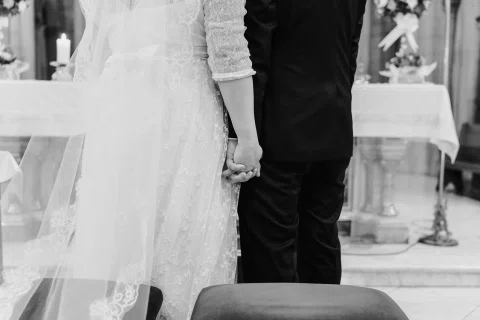 Detalle en blanco y negro de una mujer con vestido blanco y un hombre con traje oscuro tomados de la mano.