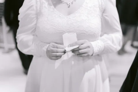 Detalle en blanco y negro de una mujer con vestido blanco sosteniendo un papel doblado entre las manos.
