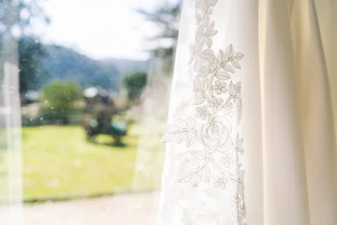 Detalle del bordado de un vestido blanco junto a una ventana con jardín al fondo.