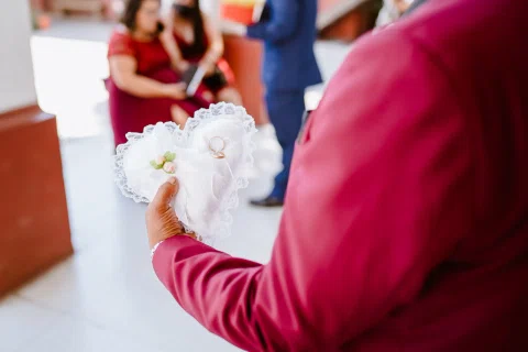 Detalle de un cojín blanco decorado con anillos, sostenido por una persona con chaqueta burdeos.