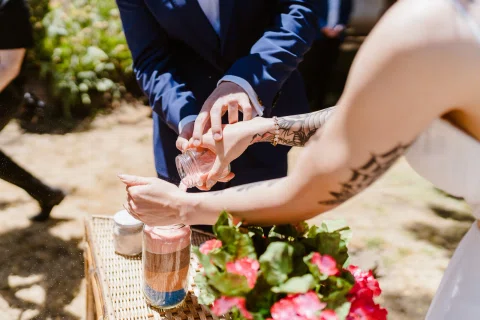 Detalle de un hombre con traje azul vertiendo material rosado desde un pequeño frasco sobre la mano de una mujer tatuada.