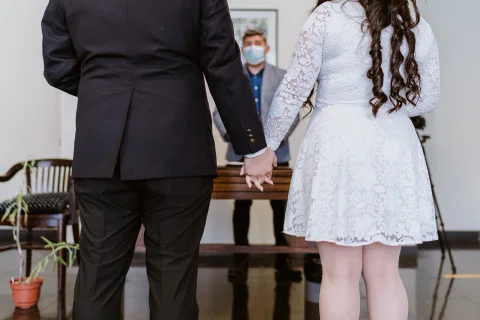 Detalle de un hombre con traje oscuro y una mujer con vestido blanco tomados de las manos, vistos desde atrás.