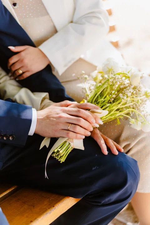 Detalle de manos con anillos junto a un ramo de flores blancas sobre el regazo de dos personas sentadas.