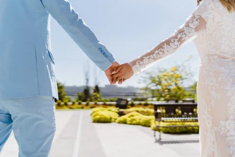 Detalle de las manos entrelazadas de un hombre con traje celeste y una mujer con vestido blanco en el exterior.