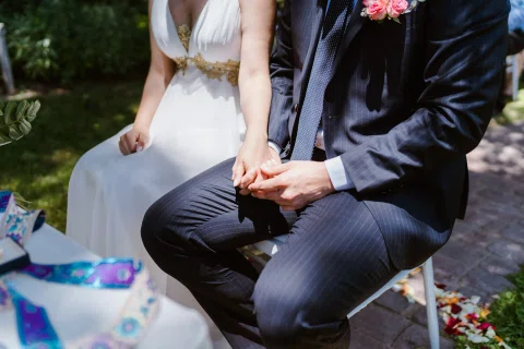 Detalle de las manos de un hombre con traje oscuro y una mujer con vestido blanco mientras están sentados.