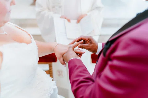Detalle de las manos de una mujer con vestido blanco y un hombre con traje burdeos frente a una persona con vestimenta blanca.