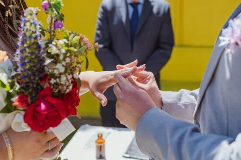 Detalle de manos mientras una persona coloca un anillo en el dedo de otra junto a un ramo de flores.
