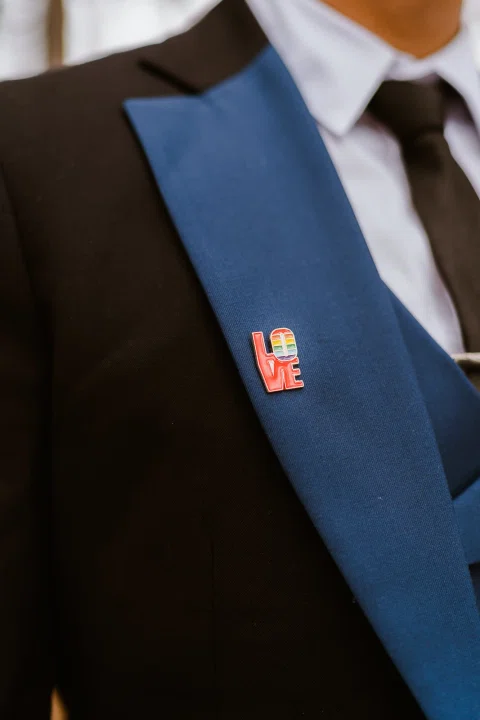 Detalle de una solapa azul sobre una chaqueta negra con un pin rojo que forma la palabra LOVE y franjas multicolor.