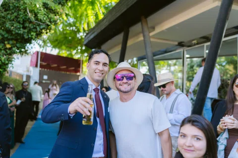 Dos hombres posan juntos al aire libre; uno viste traje azul y el otro lleva sombrero y lentes oscuros.