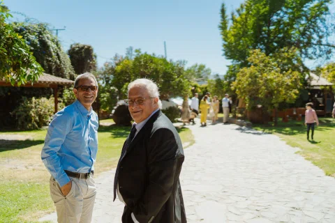 Dos hombres posan juntos en un sendero de jardín; uno viste camisa celeste y el otro traje oscuro.