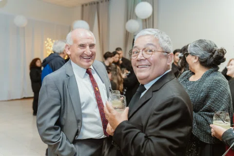 Dos hombres sonríen juntos mientras sostienen vasos dentro de un salón con más personas al fondo.