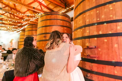 Dos mujeres se abrazan junto a un gran barril de madera dentro del salón.