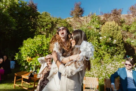 Dos mujeres se abrazan y ríen en el jardín; una sostiene un ramo blanco.