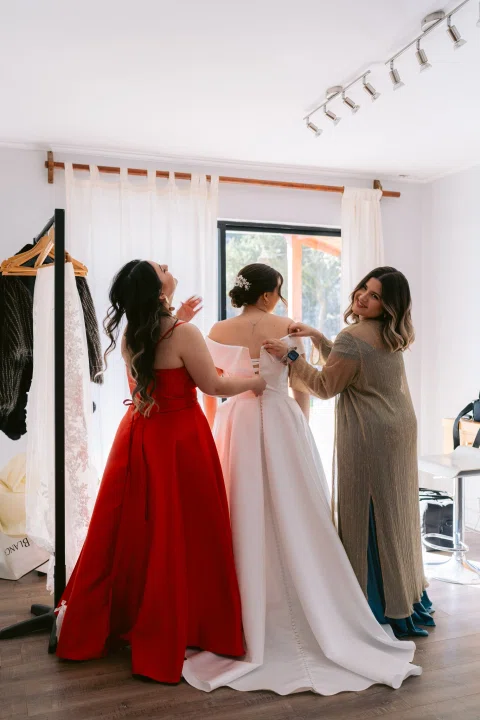 Dos mujeres ayudan a ajustar el vestido blanco de una tercera mujer dentro de una habitación.