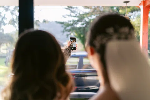 Dos mujeres vistas de espaldas miran un celular sostenido frente a ellas; una lleva velo y un automóvil aparece al fondo.