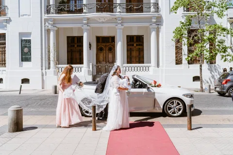 Dos mujeres de pie junto a un automóvil blanco frente a la entrada de un edificio.