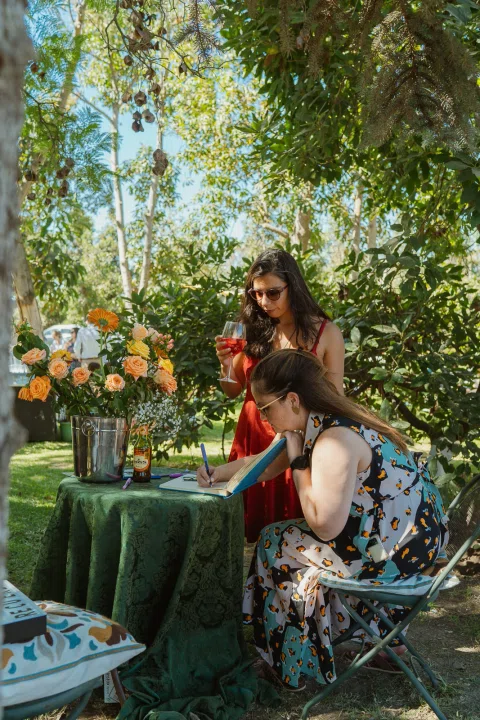 Dos mujeres permanecen junto a una mesa con flores en el jardín; una está sentada y la otra de pie.