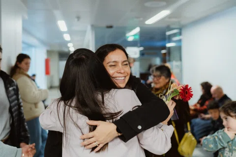 Dos mujeres se abrazan dentro de un espacio interior.