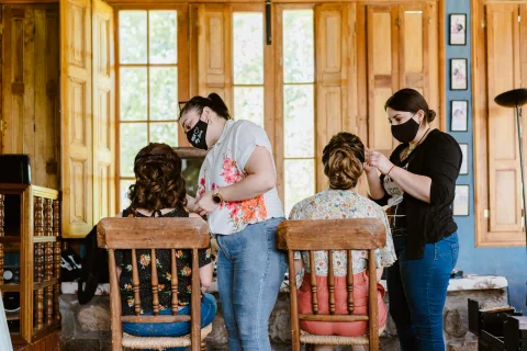 Dos mujeres sentadas mientras otras dos arreglan su cabello dentro de una habitación de madera.