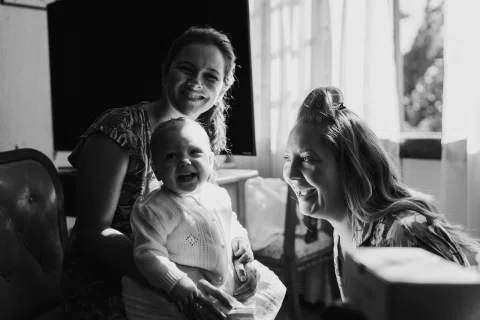 Dos mujeres sonríen junto a un bebé en una habitación, en blanco y negro.