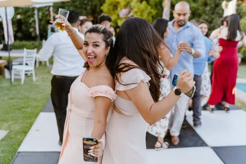 Dos mujeres con vestidos rosados bailan espalda con espalda; una levanta un vaso y ambas sostienen teléfonos.