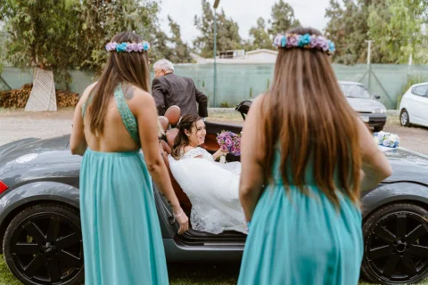 Dos mujeres con vestidos turquesa y coronas de flores caminan hacia un automóvil donde una mujer con vestido blanco está sentada.