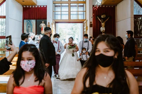 Dos niñas con mascarillas en primer plano, con una mujer de vestido blanco y un hombre de traje oscuro al fondo.