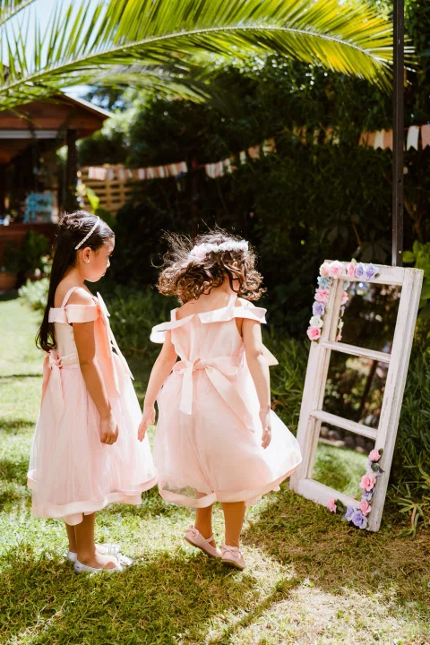 Dos niñas con vestidos rosados permanecen junto a un marco blanco decorado con flores en el jardín.