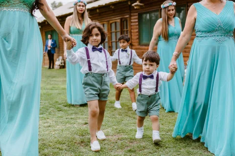 Dos niños con suspensores caminan tomados de la mano de mujeres con vestidos turquesa.