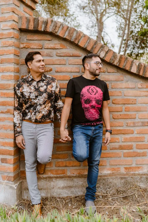 Dos personas apoyadas en un muro de ladrillo, tomadas de la mano y mirando hacia un costado.