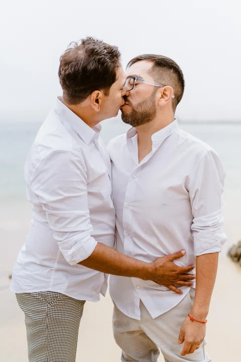 Dos personas con camisas blancas se besan en un retrato cercano frente al mar.