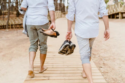 Dos personas caminan descalzas de espaldas por una pasarela de madera, cada una llevando zapatos en una mano.