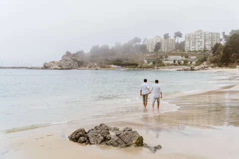 Dos personas caminan de espaldas tomadas de la mano por la orilla, con rocas en primer plano.