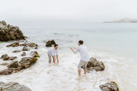 Dos personas de pie en el agua entre rocas, frente al mar en una playa.