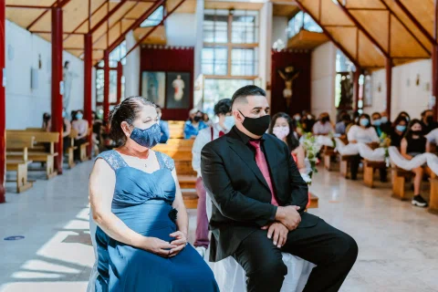 Mujer con vestido azul y hombre con traje oscuro, ambos con mascarilla, sentados dentro de una iglesia.