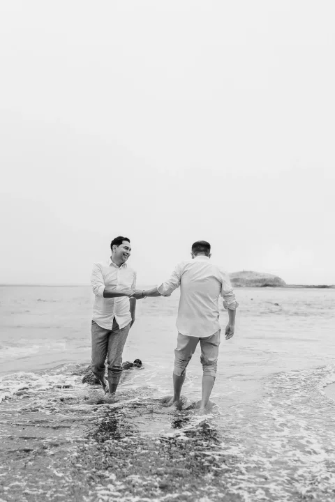 Dos personas de pie en la orilla del mar, tomadas de la mano, en una fotografía en blanco y negro.