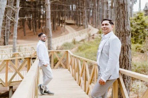 Dos personas con ropa clara están de pie a distinta distancia sobre un puente de madera entre árboles.