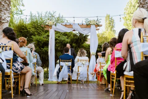 Dos personas están sentadas frente a una estructura de madera con telas blancas y flores, rodeadas por otras personas.