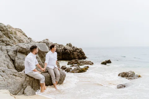 Dos personas sentadas sobre rocas junto al mar, tomadas de la mano y mirando hacia el agua.