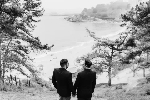 Dos personas con traje, vistas de espaldas y tomadas de la mano, frente a una bahía en una fotografía en blanco y negro.