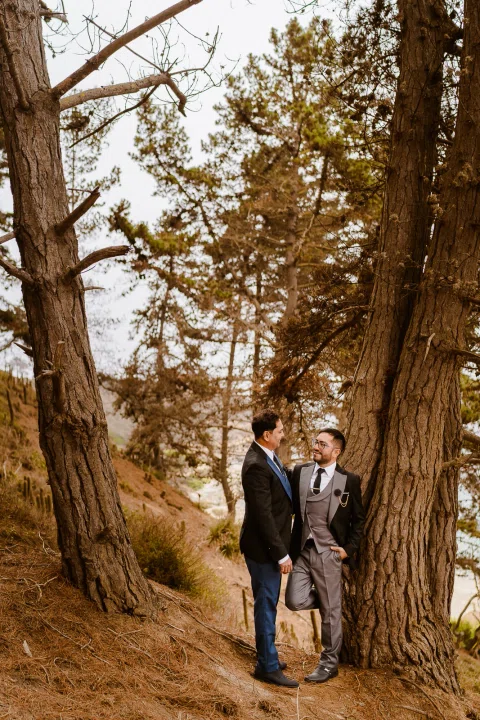 Dos personas con traje de pie junto al tronco de un árbol grande, mirándose en un entorno arbolado.