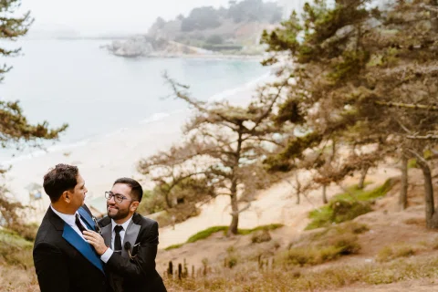Dos personas con traje frente a frente en un mirador, con una playa y el mar al fondo.