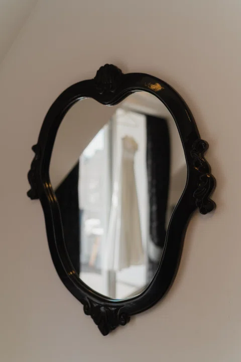 Espejo de marco negro con el reflejo desenfocado de un vestido blanco colgado junto a un ventanal.