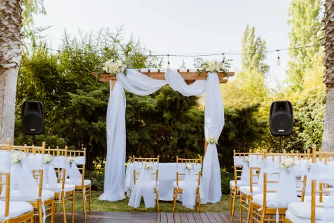 Estructura de madera cubierta con telas blancas y flores frente a filas de sillas doradas en exterior.