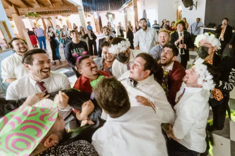 Grupo de hombres se amontona y ríe en la pista de baile, algunos con accesorios de fiesta.