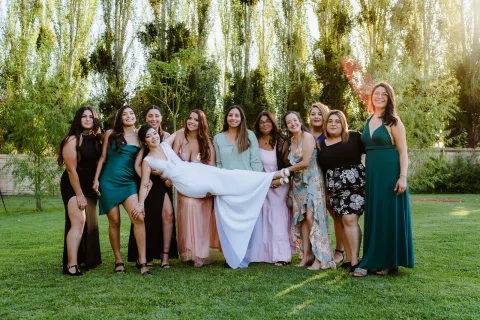 Grupo de mujeres posa en un jardín mientras varias sostienen a una persona con vestido blanco en posición horizontal.