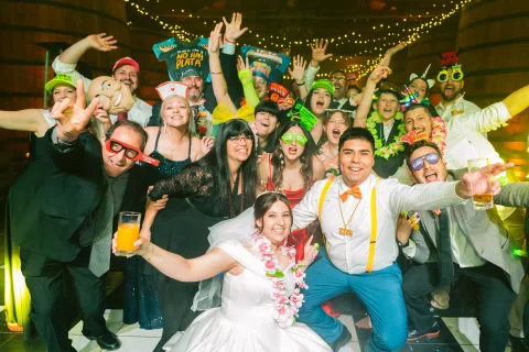 Grupo numeroso posa y celebra frente a la cámara con accesorios de fiesta y brazos levantados.