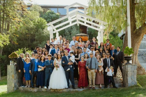 Grupo numeroso posa al aire libre junto a un hombre con traje azul y una mujer con vestido blanco.