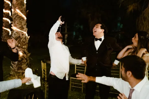 Grupo de personas celebra durante la recepción nocturna; un hombre levanta una copa mientras los demás reaccionan a su alrededor.